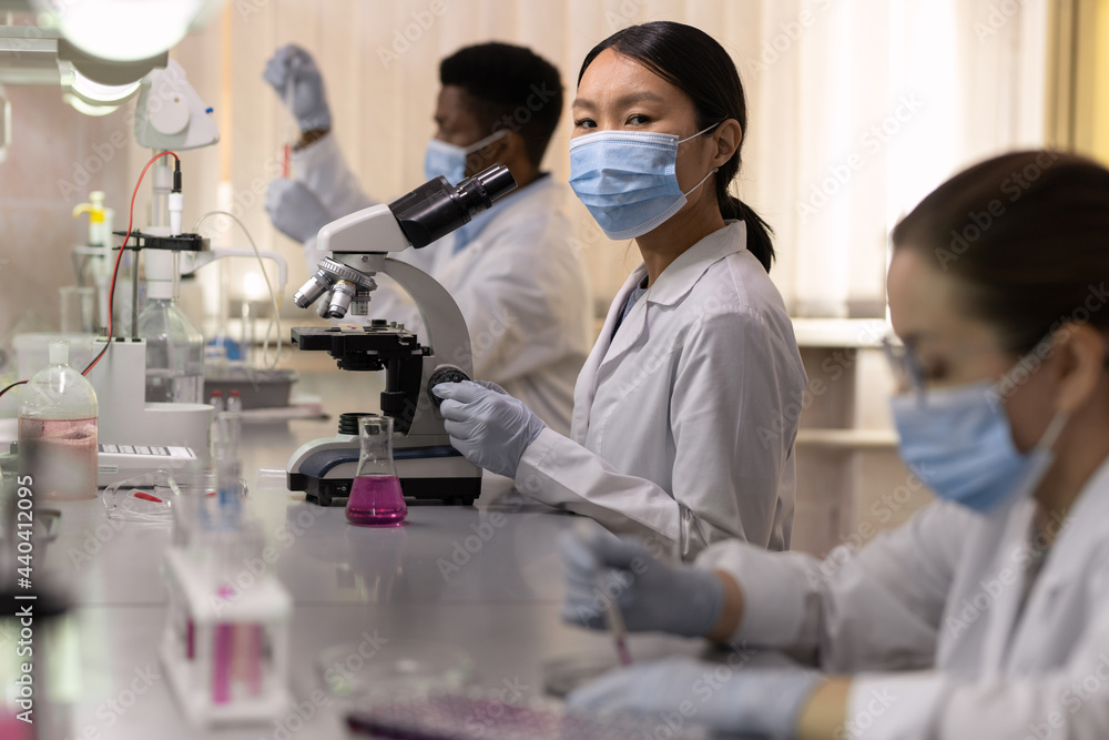Young female chemist in protective mask looking at you among colleagues Young female chemist in protective mask looking at you among colleagues