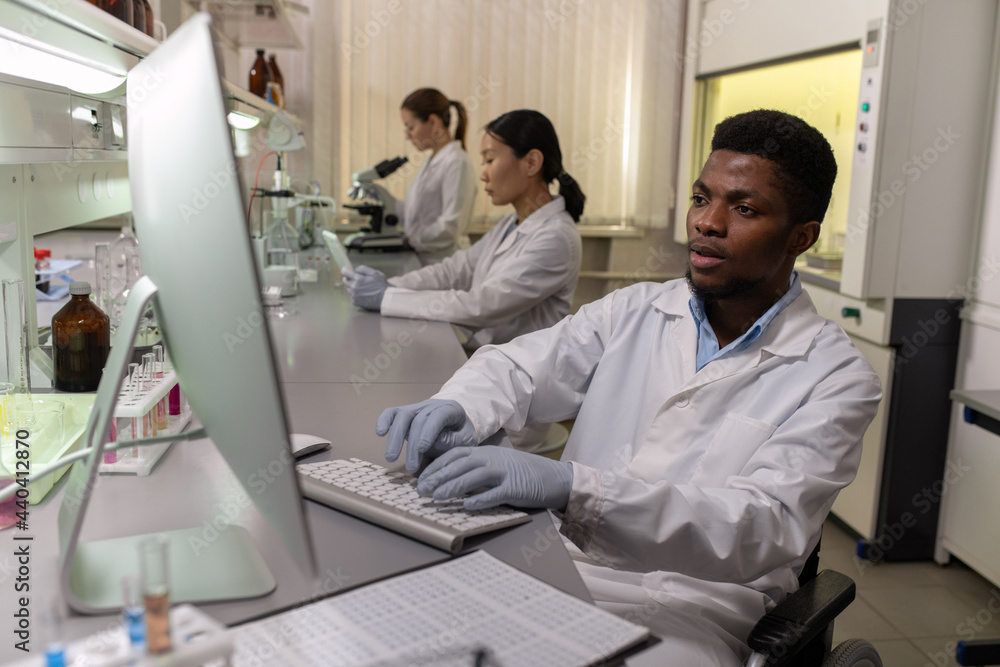 Young male scientist in gloves and whitecoat sitting in front of computer screen in lab