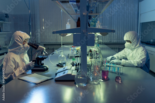 Two contemporary lab workers in coveralls using microscope and laptop