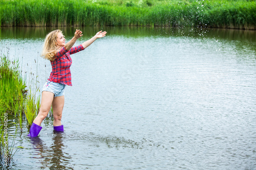 Fototapeta Naklejka Na Ścianę i Meble -  The blonde girl is having fun in the river in rubber boots in the country. Young woman on a lake enjoy the life.