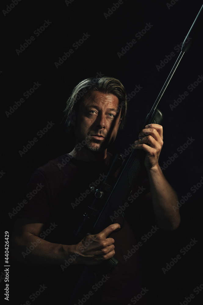 Dark portrait of blond haired man in dark brown t-shirt holding a rifle.