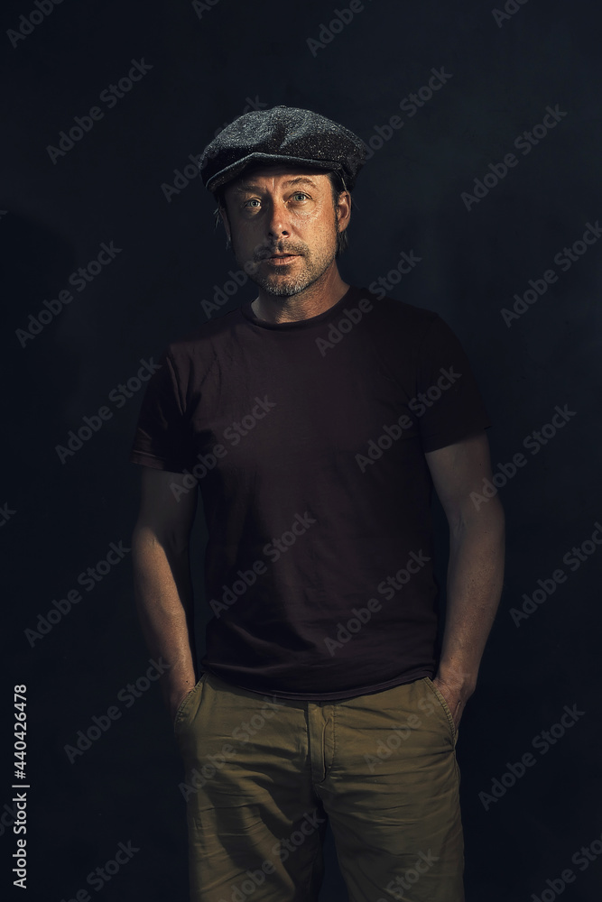 Dark portrait of a tough man with a cap and a gray stubble in a dark brown t-shirt.