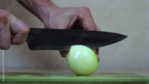 A man's hand with a knife cuts an onion into two halves on the table. Macro shot.