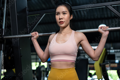 Young Asia woman lifting barbells in the fitness gym. Doing shoulder press exercise with a weight bar. Flexing muscles and making shoulder press squat. Workout and healthy lifestyle...