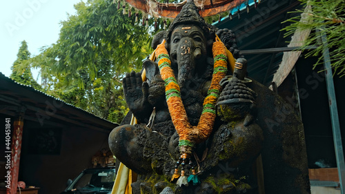 An old stone statue of Ganesha covered with moss with a wreath of yellow flowers on his neck standing on the street