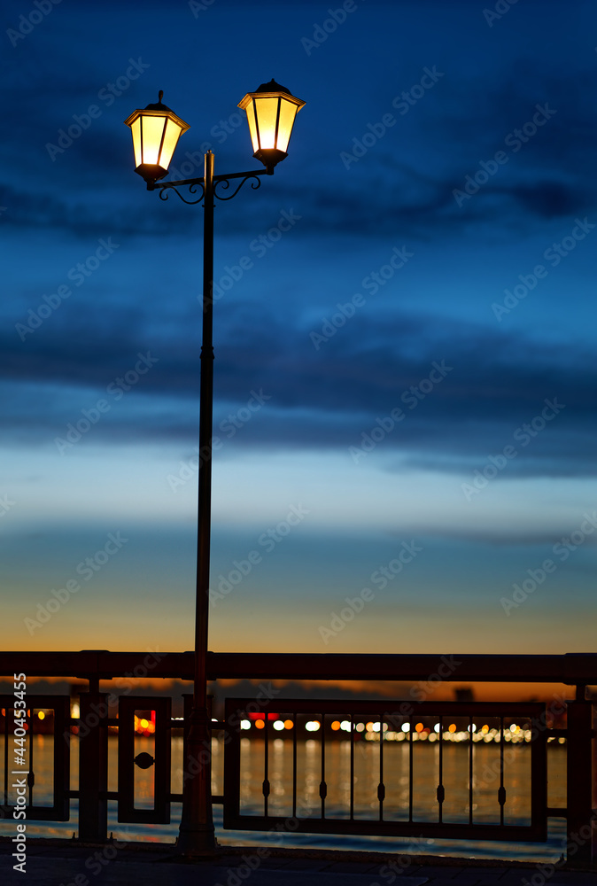 Vintage street lantern with warm yellow light and dark blue night cloudy sky on background. Night embankment river.