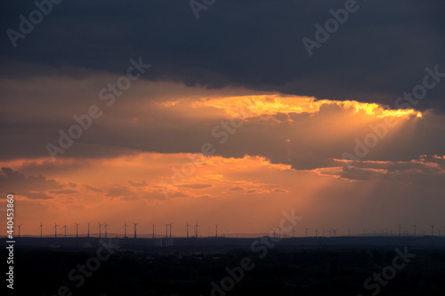 A wide landscape with many wind turbines in warm orange and red evening sunlight shining in rays through a blanket of clouds