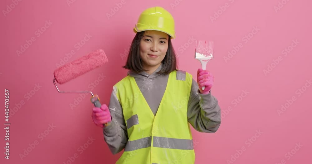Carefree positive Asian construction worker holds painting brush and roller dressed in workwear hardhat feels pleased after house refurbishment poses against pink background. Repair concept. Carefree positive Asian construction worker holds painting brush and roller dressed in workwear hardhat feels pleased after house refurbishment poses against pink background. Repair concept.