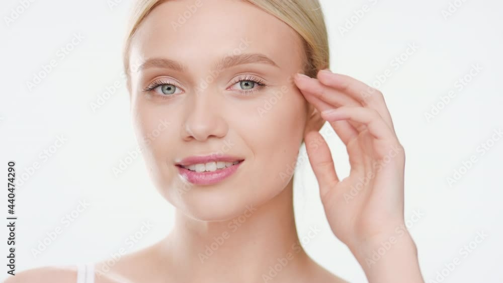 Good-looking fit European young woman with long blond hair in ponytail in white top turns to the camera touching her face and smiling wide for it on white background | Moisturizer commercial Good-looking fit European young woman with long blond hair in ponytail in white top turns to the camera touching her face and smiling wide for it on white background | Moisturizer commercial