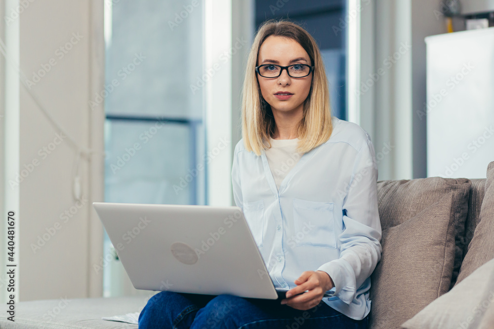 Portrait of a blonde woman looking at the camera sitting at home on the couch and holding a laptop