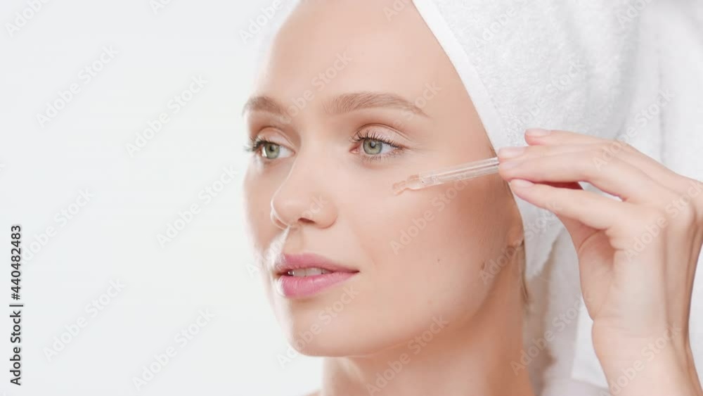 Big close-up shot of beautiful slim Caucasian young female model with towel on her hair applies face serum on her cheek from dropper on white background | Face serum commercial Big close-up shot of beautiful slim Caucasian young female model with towel on her hair applies face serum on her cheek from dropper on white background | Face serum commercial