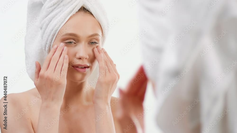 European attractive fit young woman with a towel on her head taps eye cream smiling wide in front of the mirror on white background | Eye cream commercial European attractive fit young woman with a towel on her head taps eye cream smiling wide in front of the mirror on white background | Eye cream commercial