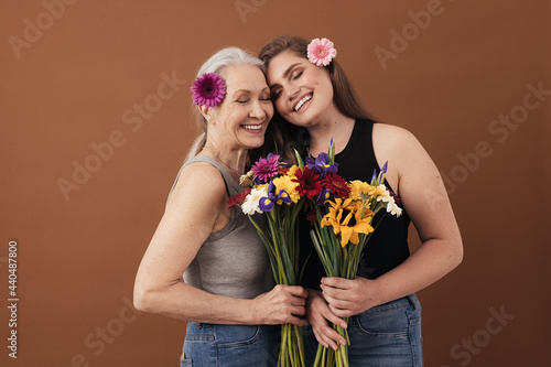 Two smiling women of different ages standing head to head in studio. Caucasian females with bouquets in hands and flowers in their hair. photo