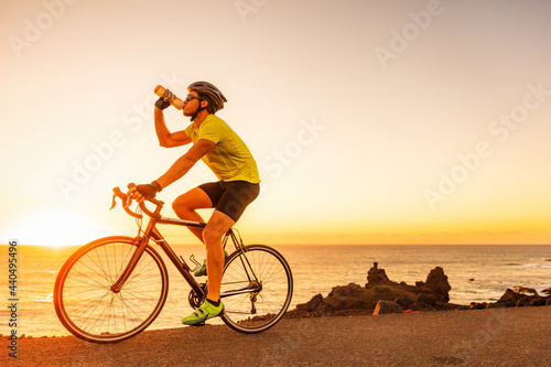 Cycling sports athlete cyclist man drinking water bottle while biking on road bike at sunset.