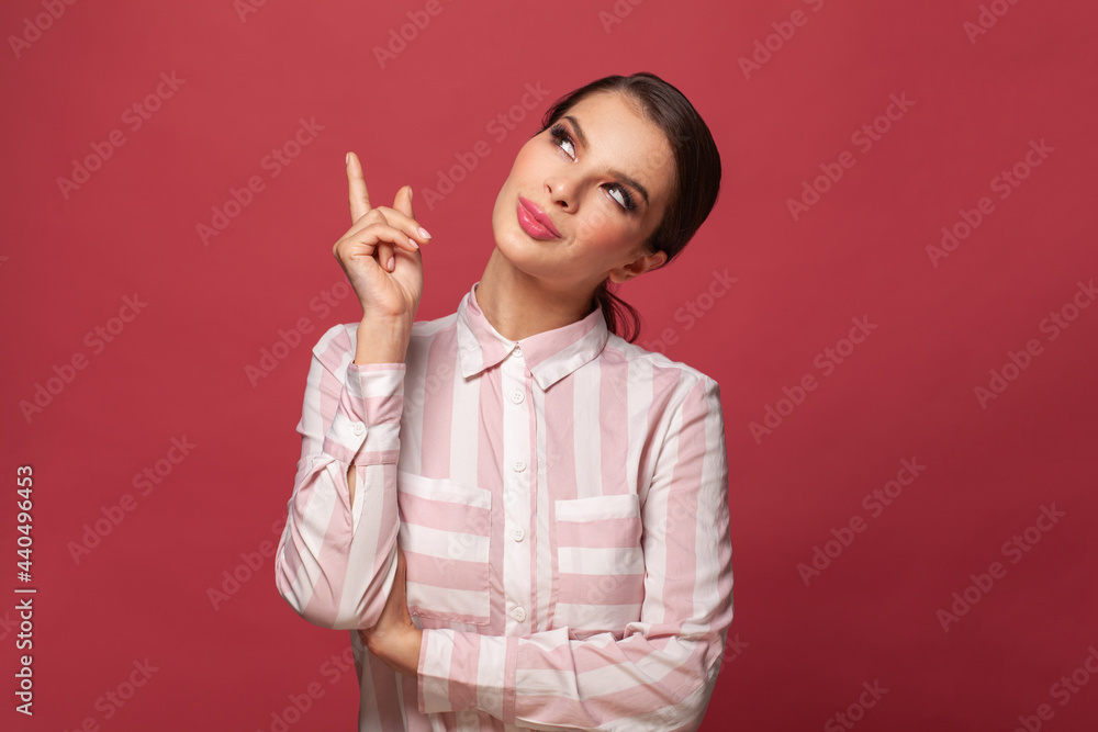 Happy smiling woman seller points up, advertises item on blank space, has friendly facial expression, wears casual shirt on red background Happy smiling woman seller points up, advertises item on blank space, has friendly facial expression, wears casual shirt on red background