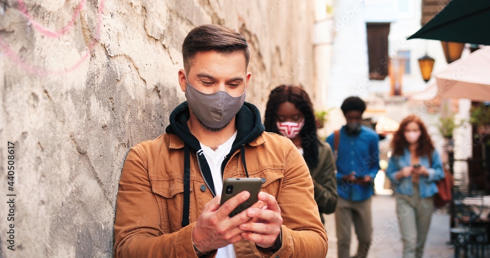 Full length view of the multiracial company of people wearing protective masks standing with each other at the safety distance and browsing internet at their gadgets while walking outdoors