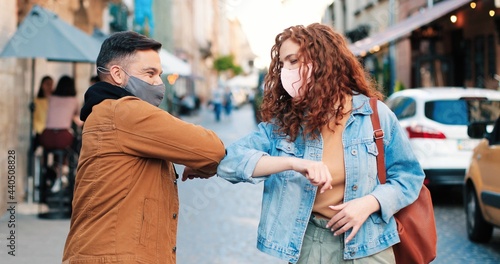 Portrait of young ginger woman bumping elbows with her caucasian male colleague as contactless greeting while walking in post pandemic period at the summer street
