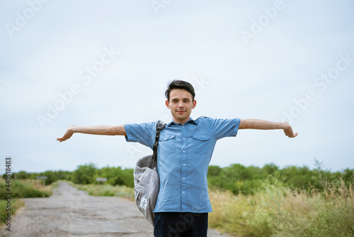 Happy young man posing with raised arms, standing on the road and looking into the distance. Happy guy in a blue shirt with a backpack. Free student enjoys vacation