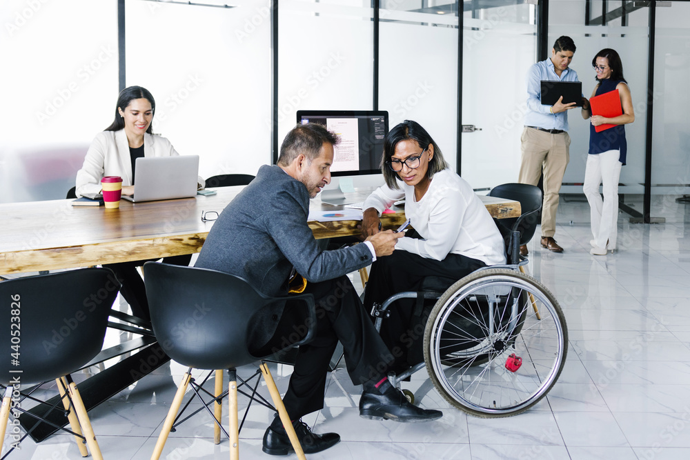 Latina transgender businesswoman in wheelchair with office colleague checking email on smartphone and office employees working in the background, in disability concept and disabled people