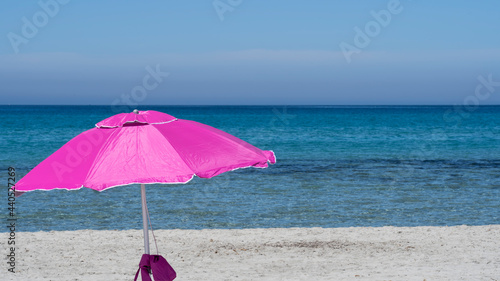 Isolated fuchsia or pink beach umbrella. Blue sky. Relaxing context. Summer holidays at the sea. General contest and location. Turquoise sea and white beach in the background © Matteo Ceruti
