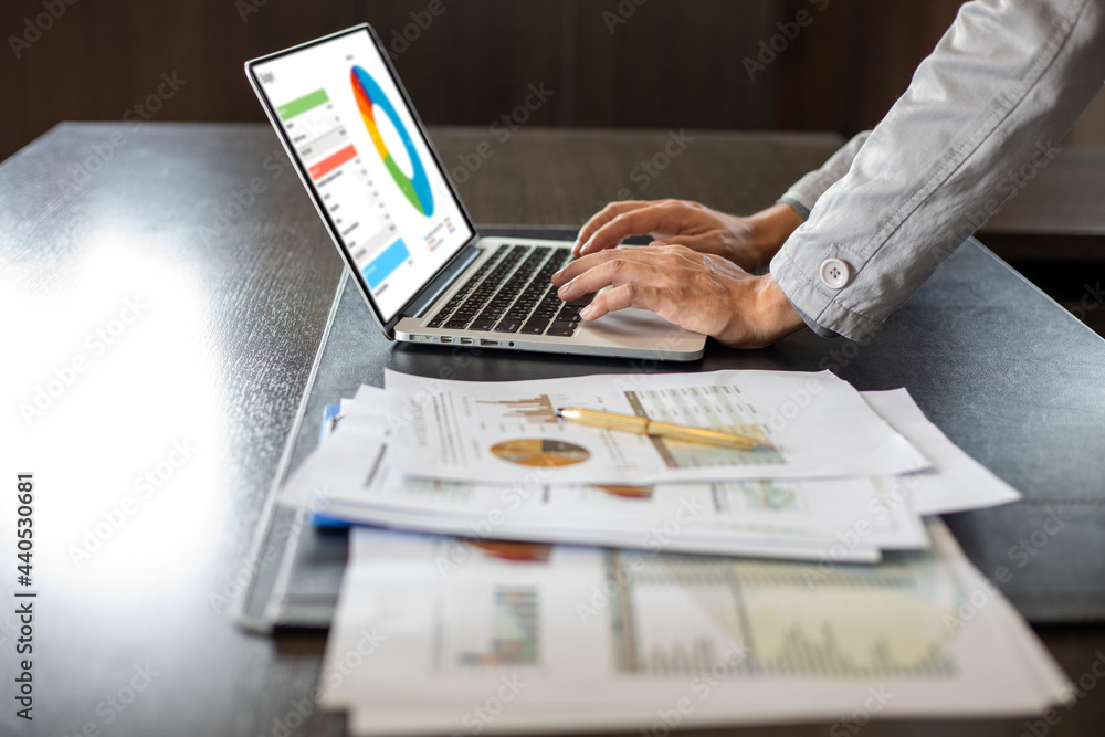 Close up Asian businessman using laptop computer analysis documents and graph financial diagram on office table working Close up Asian businessman using laptop computer analysis documents and graph financial diagram on office table working