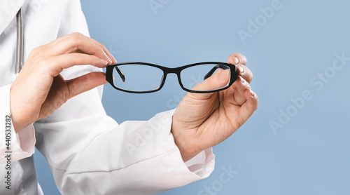 Hands of female doctor ophthalmologist or optician holds pair of glasses with black frame. Minimal banner, blue background. Copy space. Optics oculist, eye test checkups. Medicine mockup template