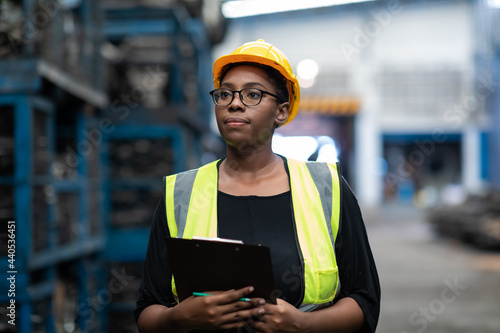 Plus size black female worker wearing safety hard hat helmet inspecting old car parts stock while working in automobile large warehouse