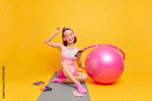 Indoor shot of carefree young Asian woman enjoys favorite playlis holds mobile phone wears stereo headphones dressed in activewear sits on mat trains at home uses fitness ball hula hoop expander