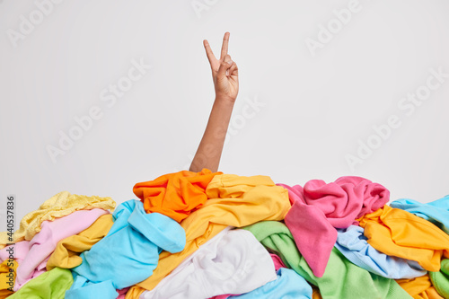Faceless individual raises arm over heap of unfolded multicolored clothes makes peace gesture isolated over white background. Unrecognizable cluttered woman. Messy laundry. Wardrobe cleaning