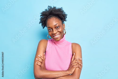 Portrait of good looking woman with dark skin curly hair smiles pleasantly keeps arms folded tilts head dressed in pink shirt isolated over blue background. Human emotions and sincere feelings concept