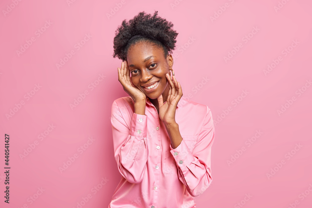 Pretty dark skinned young woman with curly combed hair touches face gently smiles toothily has pleased face expression wears stylish shirt poses against pink background. Positive emotios concept Pretty dark skinned young woman with curly combed hair touches face gently smiles toothily has pleased face expression wears stylish shirt poses against pink background. Positive emotios concept