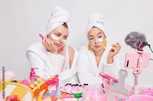 Unimpressed bored female models hold lipstick apply beauty patches under eyes shoot cosmetic product review record stylist tips vlog dressed in white bathrobes pose indoor in front of camera