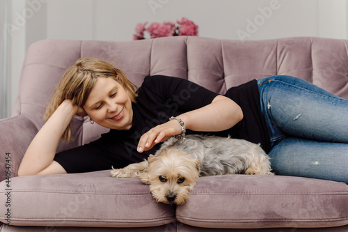 Smiling young woman is lying on pink sofa in living room, petting Yorkshire Terrier. Concept of home life. Rest and relaxation. Positive emotions.