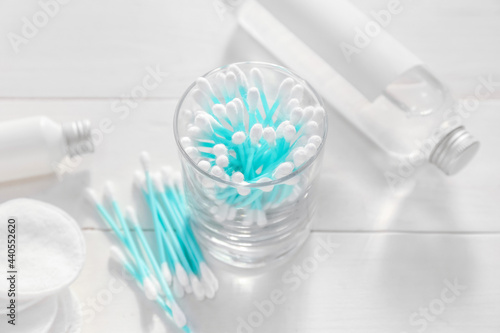 Glass with cotton swabs  pads and bottles of cosmetic products on light wooden background
