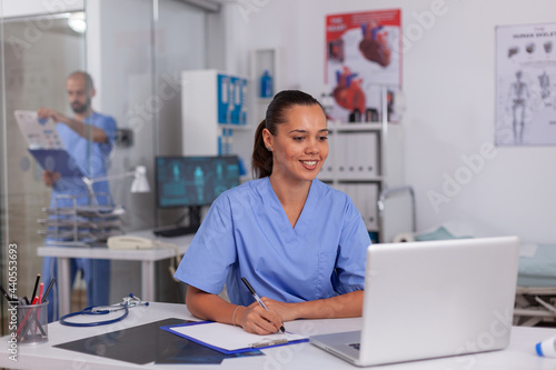 Smiling nurse using laptop computer and writing notes on clipboard in hospital office with doctor in the background. Health care physician using computer in modern clinic looking at monitor,