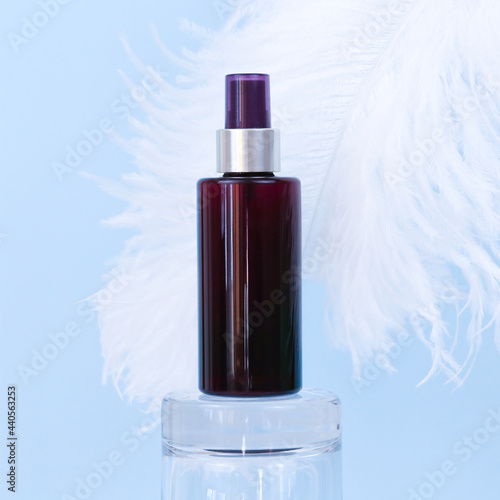 Unbranded brown sunscreen spray bottle on glass podium and large white ostrich feather on blue background. Blank label bottle for branding. photo