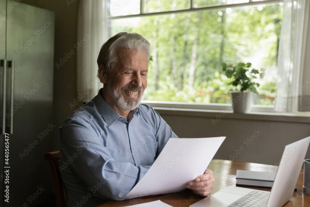 Happy old Caucasian man sit at desk at home office work on computer with paperwork documents. Smiling mature male feel excited read good news in paper letter correspondence using laptop paying.