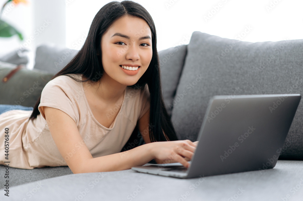 Pretty, positive chinese or japanese brunette girl lies at home on the couch on her stomach, uses a laptop, browses the Internet, chatting on social networks, looks at camera, smiles friendly Pretty, positive chinese or japanese brunette girl lies at home on the couch on her stomach, uses a laptop, browses the Internet, chatting on social networks, looks at camera, smiles friendly