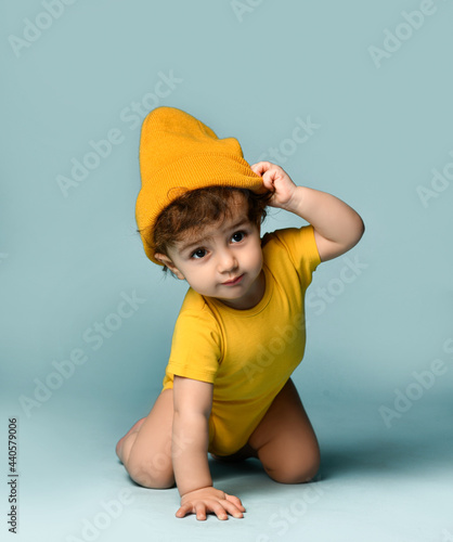Small cute smiling curly baby boy toddler in yellow comfortable jumpsuit and hat crawling on floor over blue wall background. Happy childhood, stylish look for newborn and children