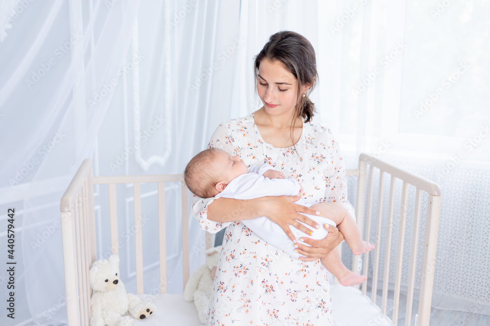 a young mother holds a newborn baby in her arms and puts it to sleep in the bedroom near the crib, the concept of motherhood and a happy family a young mother holds a newborn baby in her arms and puts it to sleep in the bedroom near the crib, the concept of motherhood and a happy family