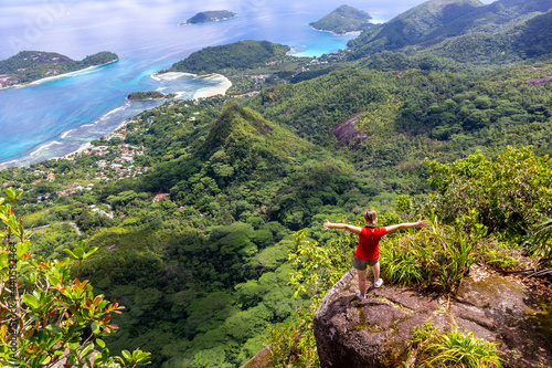 Young female traveller standing at the edge of the cliff at Morne Blanc View Point, overlooking Mahe Island coastline with lush tropical vegetation and crystal blue ocean, Seychelles.