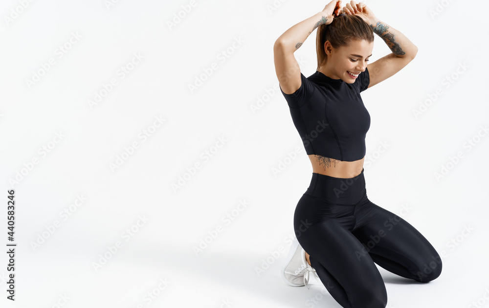 Young sport woman adjusting hair during workout, fitness girl runner prepare for exercises training session, getting ready for gym, sitting on floor in black sports clothing, white background