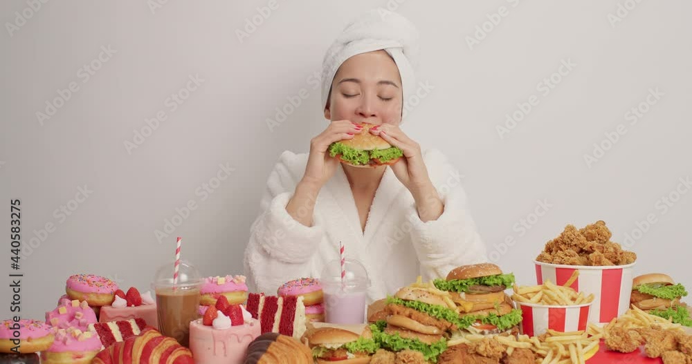 Beautiful young Asian woman feels very hungry eats tasty burger surrounded by different delicious fatty snacks wears bathrobe and towel over head isolated over white background. Cheat meal day