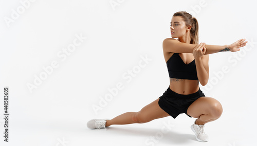 Dynamic movement. Determined young sportswoman in sport clothing, stretching legs and arms, body warm up before jogging, active athlete training session, white background © Liubov Levytska