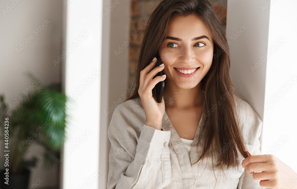 Close up portrait of smiling girl talking on mobile phone near window. Girl standing at home and having phone call, having conversation on smartphone Close up portrait of smiling girl talking on mobile phone near window. Girl standing at home and having phone call, having conversation on smartphone