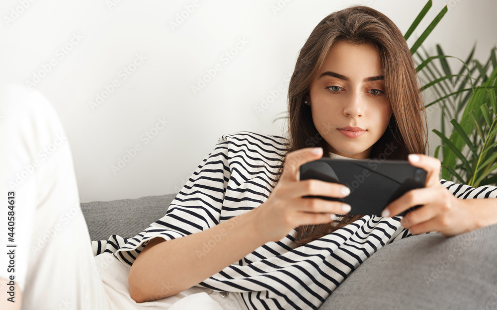 Young attractive woman lying on sofa and watching video on smartphone. Girl holding mobile phone horizontal, looking at screen, playing video game while relaxing at home Young attractive woman lying on sofa and watching video on smartphone. Girl holding mobile phone horizontal, looking at screen, playing video game while relaxing at home