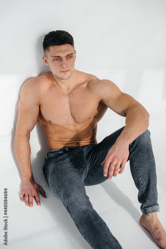 A young sexy athlete with perfect abs sits on the floor in the studio topless in jeans in the background. Healthy lifestyle, proper nutrition, training programs and nutrition for weight loss. A young sexy athlete with perfect abs sits on the floor in the studio topless in jeans in the background. Healthy lifestyle, proper nutrition, training programs and nutrition for weight loss.