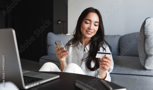 Portrait of young woman, asian girl shopping online, paying bills with credit card, laptop and smartphone, enter card number in mobile phone app to purchase smth in internet store