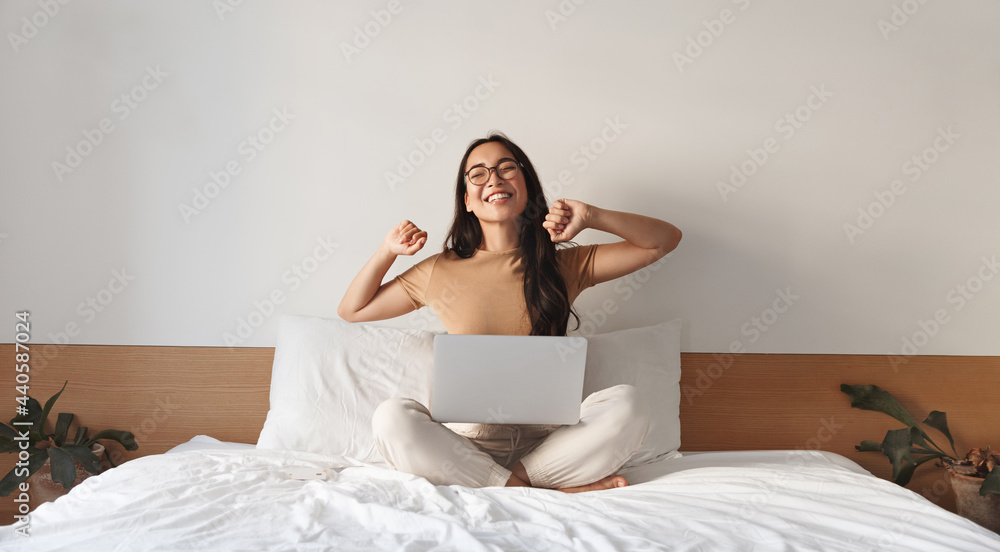 Image of satisfied young woman stretching arms, finish working on laptop. Asian girl sitting with computer in bed, wearing glasses, studying at home, e-learning and remote freelance job concept Image of satisfied young woman stretching arms, finish working on laptop. Asian girl sitting with computer in bed, wearing glasses, studying at home, e-learning and remote freelance job concept