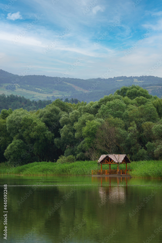 Spring natural landscape with a lake surrounded by green foliage of trees and wooden cabin Spring natural landscape with a lake surrounded by green foliage of trees and wooden cabin